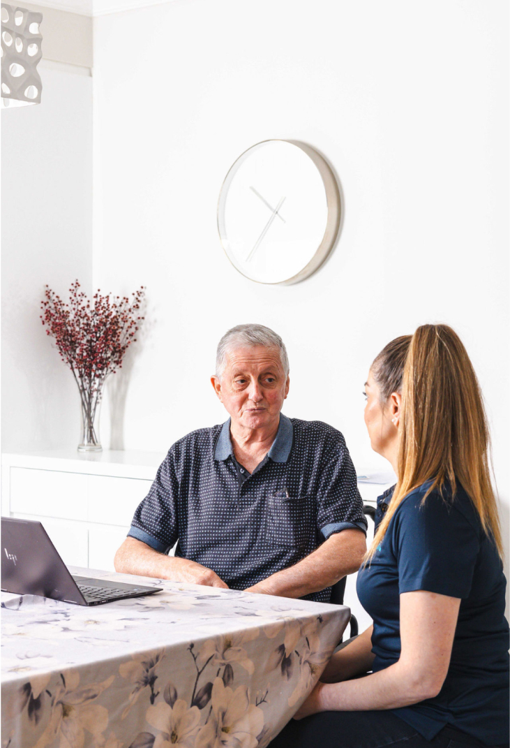 While sitting at a table, an aged care recipient has a conversation with his caregiver from Carlisle Seniors Centre.